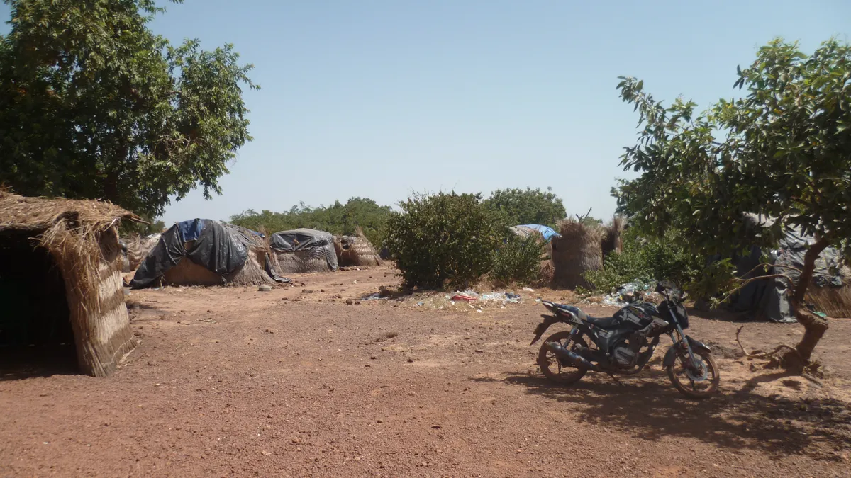 Abris de paille et de bâches d'un campement d'orpailleurs sur la latérite rouge du Burkina Faso, une moto garée à l'ombre, en 2017